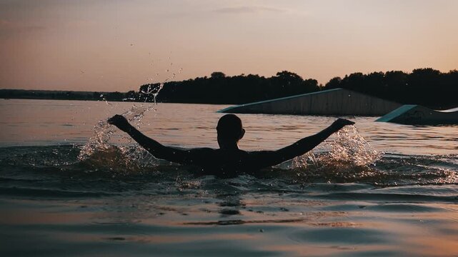 Silhouetted man emerges from a calm lake water while raising his arms in slow motion. Droplets scatter against the warm evening sky near an empty wakeboarding ramp.