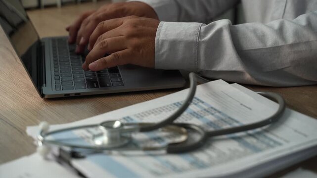 Hands typing on laptop beside stethoscope and medical paperwork, showing health insurance document review, healthcare administration and records management.