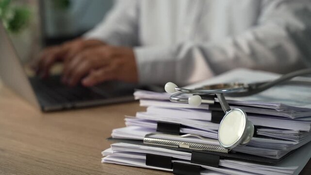 Stethoscope on stacked medical paperwork and clipboards with person working on laptop, showing healthcare documentation and medical records administration.