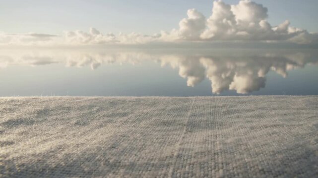Starting dolly revealing woven surface meeting mirror horizon, showing reflected cumulus and mist