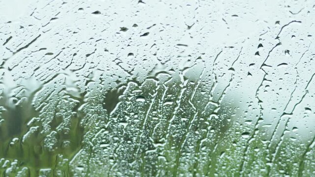 Heavy rain drops sliding down glass window with blurred green garden background in autumn weather.