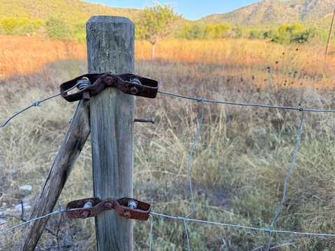 Weathered wooden fence post with rusty tensioners and barbed wire marking a property boundary across dry, sunlit fields of calvia, balearic islands countryside, copy space