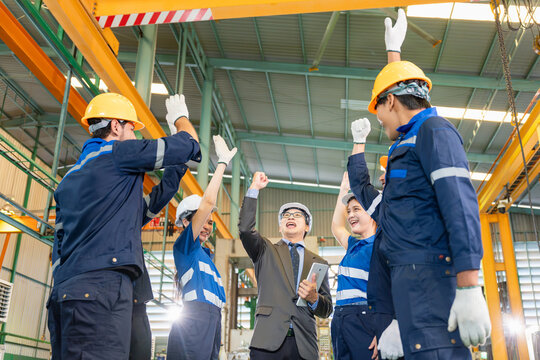 Industrial team celebrating success in factory, team of engineers and workers raising hands together in victory at factory, successful manufacturing workforce and managers celebrating inside a plant