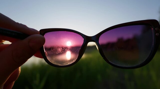 Hand holds tinted sunglasses framing the vibrant sunset reflection across a tranquil lake. Soft evening backlighting evokes nostalgic summer memories for lifestyle travel storytelling.