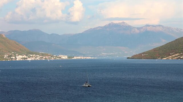 Timelapse of clouds moving over a scenic Adriatic bay with a sailing boat on blue water, coastal towns and high mountains in the background.