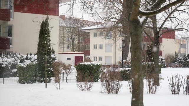 Snowy residential yard in Germany shows modern apartment buildings, balconies, hedges and bare trees during winter snowfall. Quiet urban neighborhood reflects cold weather, housing and seasonal life