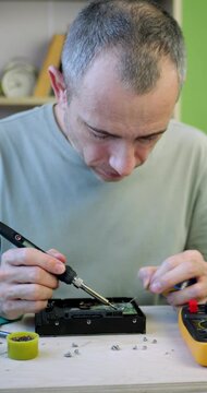 Man solders hard drive circuit board in home workshop using precision tools. Vertical scene highlights electronics repair, technical expertise and computer hardware maintenance.
