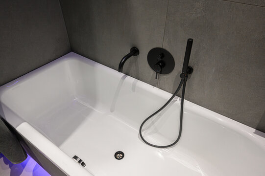 Sleek white bathtub with matte black wall-mounted fixtures, minimalist design, gray microcement walls, handheld showerhead, clean lines, modern aesthetic