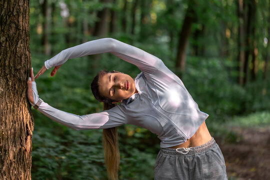 Woman doing a side stretch beside a tree. Forest fitness and flexibility exercise.