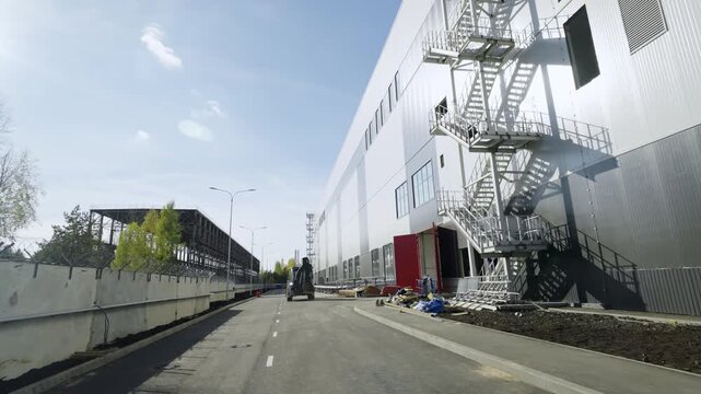 Durable and sturdy emergency staircases attached to facade of extensive warehouse. Powerful forklift moves steadily along broad paved road