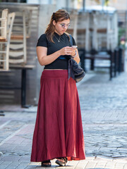 Obraz premium Teenage girl wearing a long red skirt looking intently at a smartphone on the city streets