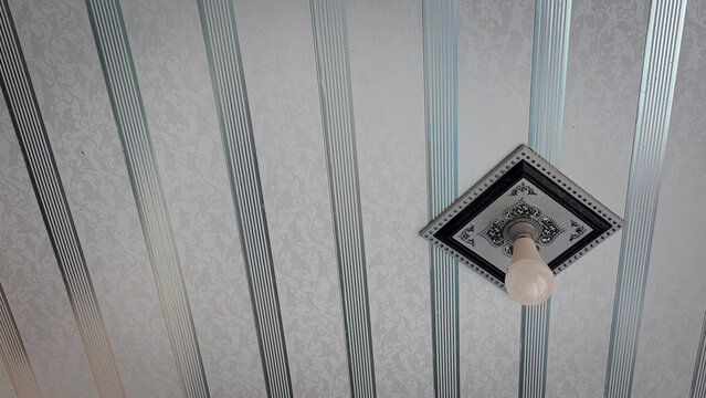 Modern LED light bulb on a decorative square base against a striped PVC panel ceiling interior.