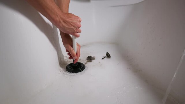 Strong hands operate a black rubber suction plunger over an open tub drain. A removed stopper and accumulated wet hair debris lie on the white ceramic surface beside pooled water.