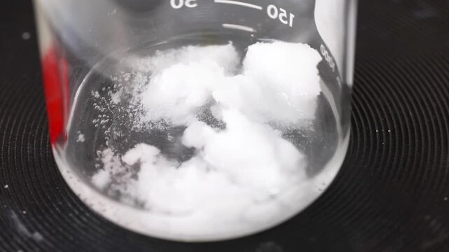 Clear liquid and white solid substances bubbling in a glass beaker, undergoing chemical reaction on a black surface.