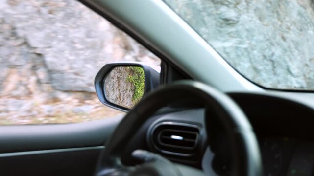 Driving Along Narrow Mountain Road from Inside Car. View from inside a moving car driving along a narrow mountain road beside a stone wall, with side mirror and steering wheel visible.