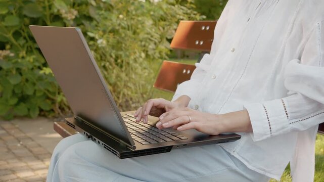 A woman is typing on a laptop in an outdoor area while sitting on a bench. The green plants surround her as she focuses on her work under natural light.