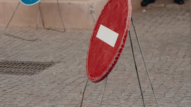 A red and white no entry sign stands on a cobblestone street in Morocco. People walk by, unaware of the restriction. The sign is weathered, adding to the charm of the scene.