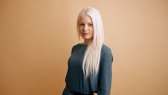 Young woman posing in studio, confident expression, long blonde hair, wearing a green ribbed sweater, beige background