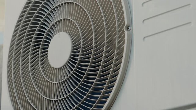 Close-up of a white air conditioning unit fan spinning. The fan blades create a hypnotic swirling effect. The unit is mounted on a wall and provides cool air.