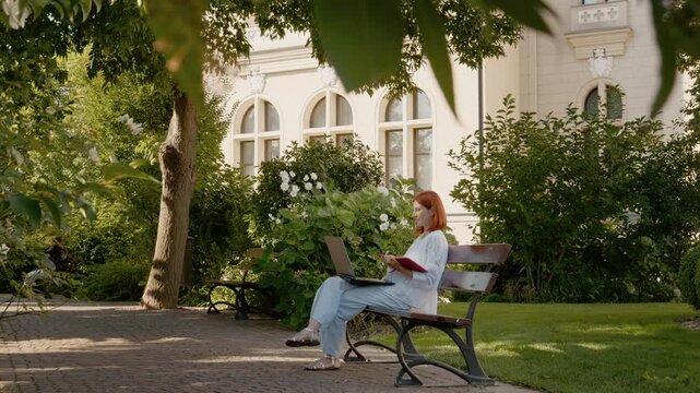A woman is sitting on a bench outside while having a video call on her laptop. She holds a small notebook and engages in conversation in a garden setting.