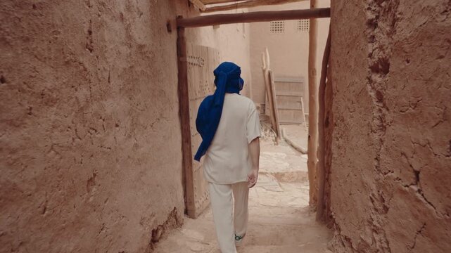 A man walks through the narrow streets of a Moroccan village. The walls are made of mud and the streets are dusty. He's wearing traditional Moroccan clothing and a blue turban.