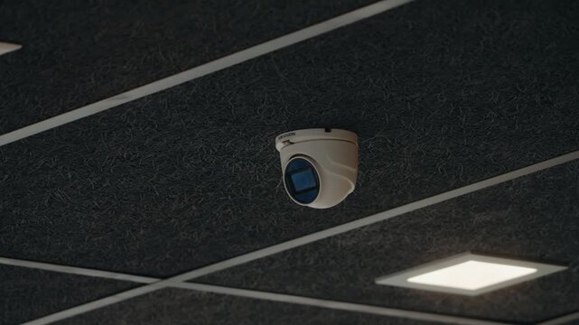 A white Hikvision CCTV security camera is mounted on a dark ceiling. The camera lens is facing downwards. The ceiling is made of square panels. Two bright LED panel lights provide illumination.