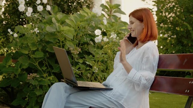 A woman sits on a bench outside, talking on the phone and typing on her laptop. She continues her work while engaged in conversation among the flowers.