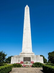Fototapeta premium White stone obelisk war memorial against a clear blue sky with red poppy wreaths placed at the base