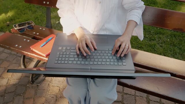 A woman sits on a bench outside in a park. She opens her laptop and starts to type. The setting is bright with grass and trees around her.