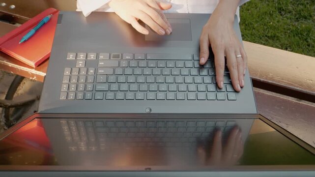 A female student is focused on her laptop while sitting on a bench outside. She uses her finger to navigate the touchpad. A notebook is placed beside her.