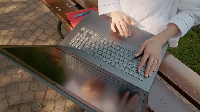 A female student is sitting on an outdoor bench in a park. She is working on a laptop and moving her finger across the touchpad.