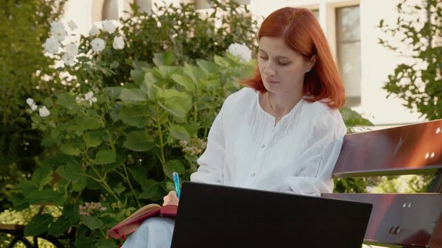 A female student sits on a bench outside, using her laptop to join a video call while taking notes in a small notebook.