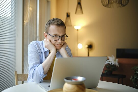A middle-aged man with glasses watching series on his laptop while seated at a round table in a stylish home. Natural light fills the space, creating a calming atmosphere for productivity