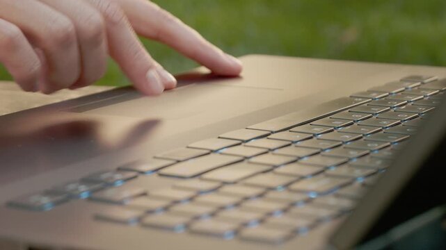A woman fingers move across the touchpad of a laptop, then press down on a key. The setting appears to be outside, with grass in view.