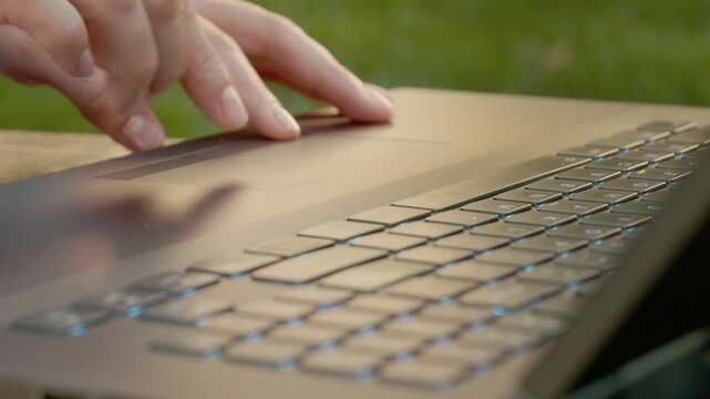 A woman navigates the touchpad of a laptop in a park. The focus is on her fingers as they move on the device while the sun shines around her.