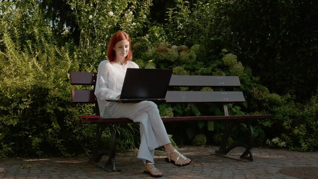 A female student is sitting on a bench and using her laptop in a garden outside. She appears focused on her work while surrounded by greenery and plants.