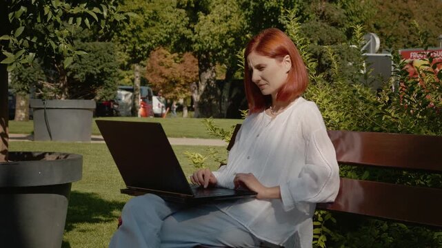 A female student is sitting on a bench outside in a garden. She is frowning and focused on her laptop. The setting is bright with greenery around her.