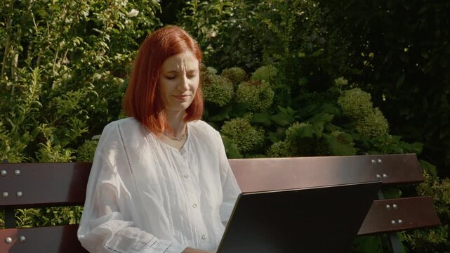 A female student sits on a bench in a garden. She appears tired and takes a deep breath while looking at her laptop screen and continues her work.