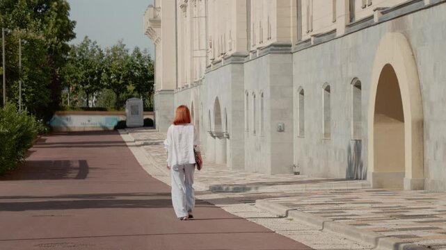 A female student walks along the path past a university building holding a laptop. She is on her way to class. It is a clear day with trees nearby.