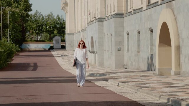 A female student walks along a path by a university building, carrying her laptop. She moves towards the camera on a clear day.