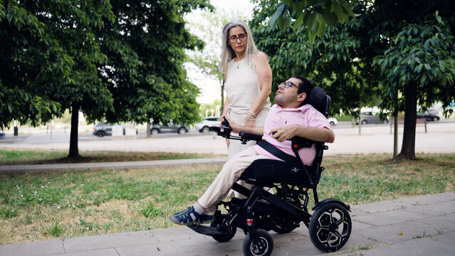 Man with spina bifida navigating a park path in a motorized wheelchair, accompanied by a caregiver, focusing on inclusion and support