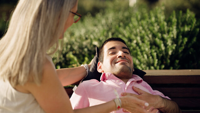 Caregiver providing comfort and support to a man with spina bifida, resting on a park bench during a sunny day