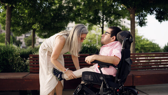 Caregiver assisting a man using a wheelchair, gently stretching his leg in a green park setting, promoting health and wellness