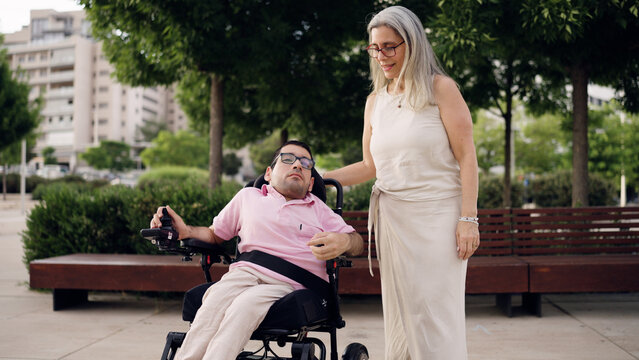Man with spina bifida in electric wheelchair getting assistance from a woman caregiver in a park