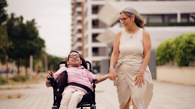 Happy disabled man in wheelchair holding hands with woman, walking together outdoors. Representing care, support, and friendship