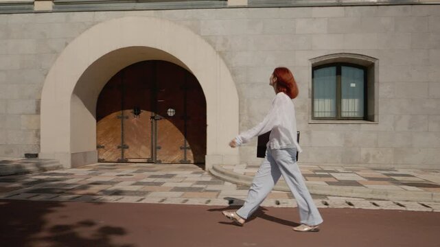 A female student carries a laptop and walks along a path next to a university building. The sun shines as she moves forward on campus.