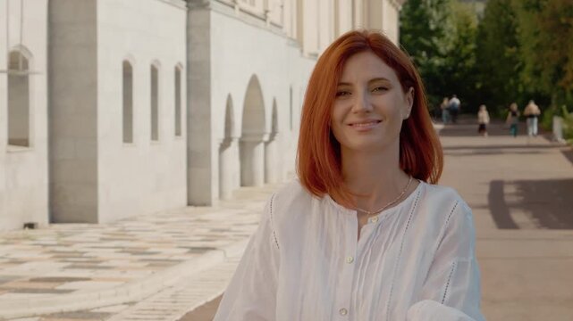 A red-haired woman walks through a park. She turns and looks at the camera for a moment. The park has trees and people walking in the background.