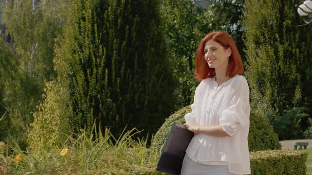 A female student walks through a park with her laptop in her arms. The sun is shining and green trees surround her. She seems focused and purposeful.
