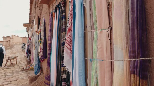 Colorful scarves and textiles hang on a building in a Moroccan market. A person walks in the background. The market offers a variety of traditional Moroccan goods.