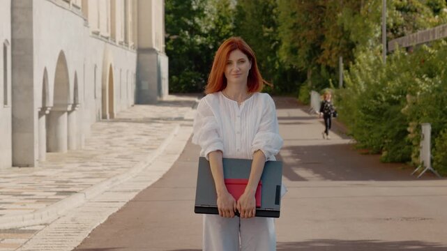 Redhead woman stands outside while holding a laptop. She looks at the camera with a serious expression. A pathway and green trees are visible in the background.